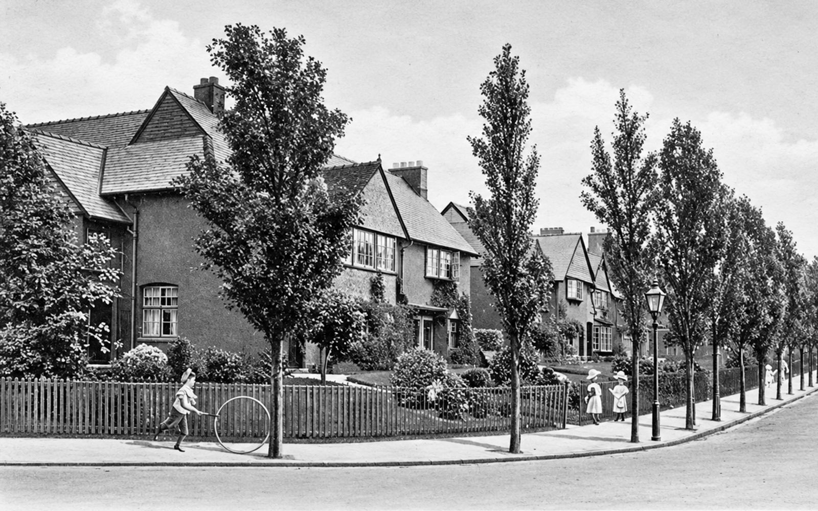 A tree-lined village street, with a boy bowling a hoop past the cottage gardens
